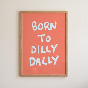 May include: A rectangular wall art print in a light wood frame featuring a coral-colored background with the hand-lettered text in light blue that reads BORN TO DILLY DALLY. The lettering has a casual, brush-stroke style against the solid color.