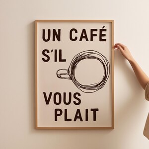 以下が含まれることがあります： 白い背景に黒い文字で「Un café s'il vous plaît」と書かれた額装されたプリント。テキストの上にコーヒーカップのシンプルな線画が中央に配置されています。