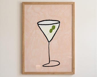 May include: A framed wall art print featuring a minimalist black line drawing of a martini glass with two green olives inside. The background is a soft, textured pale pink color, and the artwork is encased in a light-toned wooden frame.