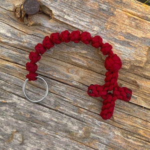 May include: A red knotted rope keychain with a cross charm and a silver ring. The keychain is lying on a weathered wooden surface.