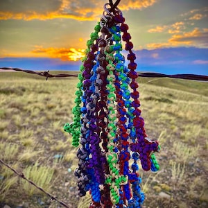 May include: A collection of colorful braided rope bracelets with a cross charm hanging from a metal ring. The bracelets are hanging on a barbed wire fence against a sunset sky.
