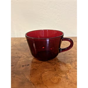 May include: A translucent ruby red glass cup featuring a smooth, rounded bowl shape and a small, curved handle on the side. The cup has a deep, consistent crimson color throughout the glass material, with a simple, functional design and a wide rim.
