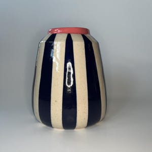 Puede incluir: Un jarrón de cerámica con cuerpo redondeado y parte superior estrecha. El jarrón presenta rayas verticales en azul marino y crema, con un borde de color coral. El diseño es moderno y el jarrón es adecuado para la decoración del hogar.