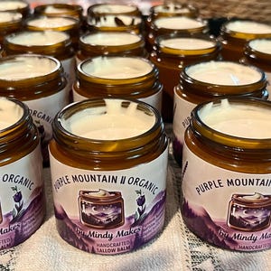 Op de afbeelding: Meerdere amberkleurige glazen potten gevuld met witte talkbalsem, elk voorzien van een etiket met de tekst Purple Mountain Organic by Mindy Makes, met een illustratie van een berglandschap en lavendeltakjes op een witte achtergrond.