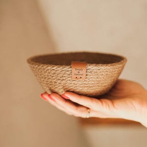 Może przedstawiać: Tkana miska z juty w naturalnym kolorze. Miska jest trzymana w dłoni i posiada małą, brązową skórzaną metkę z napisem "Simply & Natural". Dekoracyjny przedmiot.