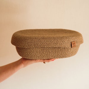 Può includere: Una mano tiene un grande cesto ovale intrecciato con fibre naturali. Il cesto ha un'etichetta in pelle marrone con il testo "Simply Natural".