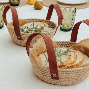 May include: Two brown woven baskets with leather handles filled with sliced bread and rosemary sprigs. The baskets are on a white tablecloth.