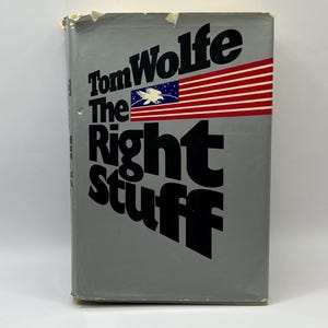 Puede incluir: Un libro de tapa dura gris con el título "The Right Stuff" de Tom Wolfe. La portada presenta una bandera estadounidense estilizada con un águila.