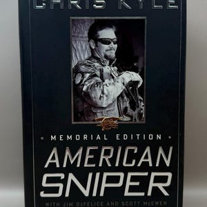 May include: A hardcover book titled "American Sniper" by Chris Kyle, with Jim DeFelice and Scott McEwen. The cover features a black and white photo of a man in military attire and the text "Memorial Edition".