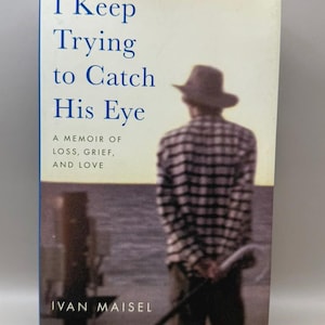 May include: A hardcover book titled I Keep Trying to Catch His Eye: A Memoir of Loss, Grief, and Love by Ivan Maisel. The cover features a person wearing a hat and a plaid shirt, standing with a cane while looking out over a body of water.