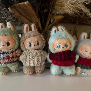 May include: Four plush dolls with bunny ear hats and knitted sweaters. The dolls have round faces with large eyes and small mouths. The sweaters are in various colors, including green, beige, red, and gray. The dolls are standing on a white surface.