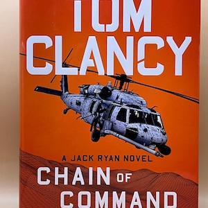 Peut inclure: Une couverture de livre avec un fond orange sur laquelle figure le titre "Chain of Command" de Tom Clancy, un roman de Jack Ryan. La couverture présente également une illustration en noir et blanc d'un hélicoptère.