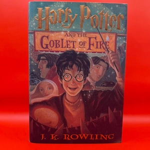 May include: A hardback book titled Harry Potter and the Goblet of Fire by J.K. Rowling. The cover features a central illustration of a young person with round glasses, surrounded by other characters, a wand, and a starry background with gold text.