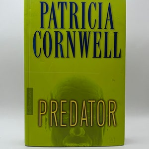 Puede incluir: Una cubierta de libro verde con el título "PREDATOR" en letras amarillas. El nombre del autor, "PATRICIA CORNWELL", está impreso en letras azules sobre el título. Una imagen en blanco y negro de la cara de una persona es visible detrás del título.