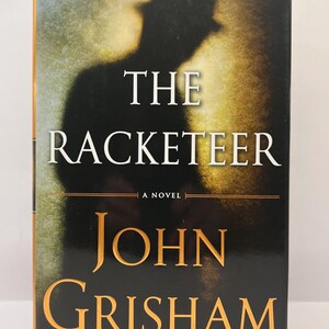 May include: A black and gold book cover with the title "The Racketeer" and the author's name "John Grisham" in a large font.