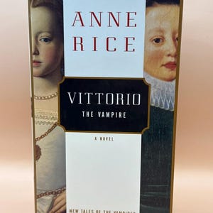 以下が含まれることがあります： アン・ライス著『Vittorio the Vampire』のハードカバー本。表紙の中央には白いパネルがあり、赤い文字で著者名が、黒い長方形のラベルにタイトルが記されている。中央のテキストの両側には、二人の人物の肖像画が配置されている。