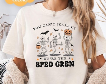 May include: A white t-shirt with the text "You can't scare us, we're the sped crew" and a graphic of three skeletons holding a pumpkin and a bucket of candy. The skeletons are wearing a black and white striped pattern. The text is in black and the graphic is in orange, black, and white.