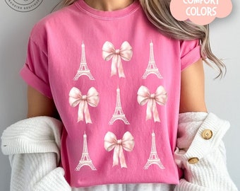May include: A bright pink short-sleeved t-shirt featuring a repeating graphic pattern of white Eiffel Tower illustrations and light pink satin-style bows arranged in a grid. The shirt has a crew neckline and rolled sleeves, with the text CRUNCHBERRY.