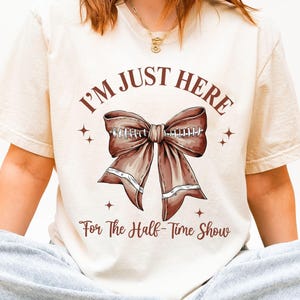May include: Ivory-colored t-shirt with the text "I'M JUST HERE For The Half-Time Show" and a brown bow graphic. The shirt also has the text "IVORY" at the bottom right.