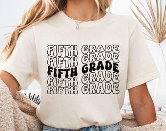Puede incluir: Una camiseta de manga corta color crema con el texto FIFTH GRADE repetido en una fuente negra ondulada y apilada. La camiseta se combina con pantalones cortos de mezclilla azul claro y un cárdigan beige texturizado para un estilo casual.