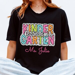 May include: Black t-shirt with the words "KINDER Assistant GARDEN Ms. Julie" in colorful, playful lettering. The design includes a polka dot border. The shirt is a casual style.