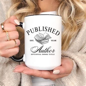 May include: White ceramic mug with black handle and rim, featuring the text "PUBLISHED Author" with an illustration of books. The mug also includes the text "EST. YEAR" and "OPTIONAL BOOK TITLE". The Quill Cottage logo is visible.