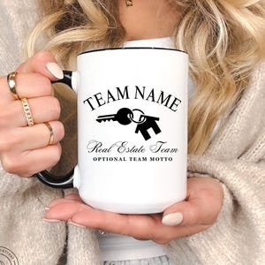 May include: White ceramic mug with black trim, featuring the text "TEAM NAME" above a key and house graphic, and "Real Estate Team" below. The mug also includes the text "OPTIONAL TEAM MOTTO".
