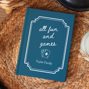 May include: A rectangular teal book with a white decorative border. The text reads all fun and games with an illustration of three playing cards below it. The bottom of the cover features the text Taylor Family in a white serif font.