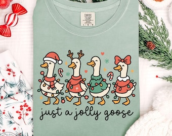 May include: A light green Comfort Colors t-shirt featuring a whimsical design of four geese dressed in festive holiday attire. Each goose is adorned with a Santa hat, reindeer antlers, or a bow, and string lights. The text "just a jolly goose" is below.