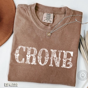 May include: Espresso-colored t-shirt with the word "CRONE" in distressed white lettering. A silver necklace with a pendant rests on the shirt. The shirt has a crew neck and a Comfort Colors tag.