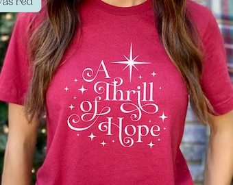 May include: A canvas red t-shirt with the text "A Thrill of Hope" in white script, accented by a star and smaller starbursts. The shirt is a solid color and has a crew neck.