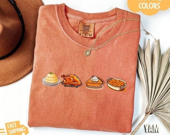 May include: A burnt orange t-shirt with a graphic of Thanksgiving dinner foods. The graphic includes mashed potatoes, a roasted turkey, pumpkin pie, and green beans. The text "Comfort Colors" is on the shirt.