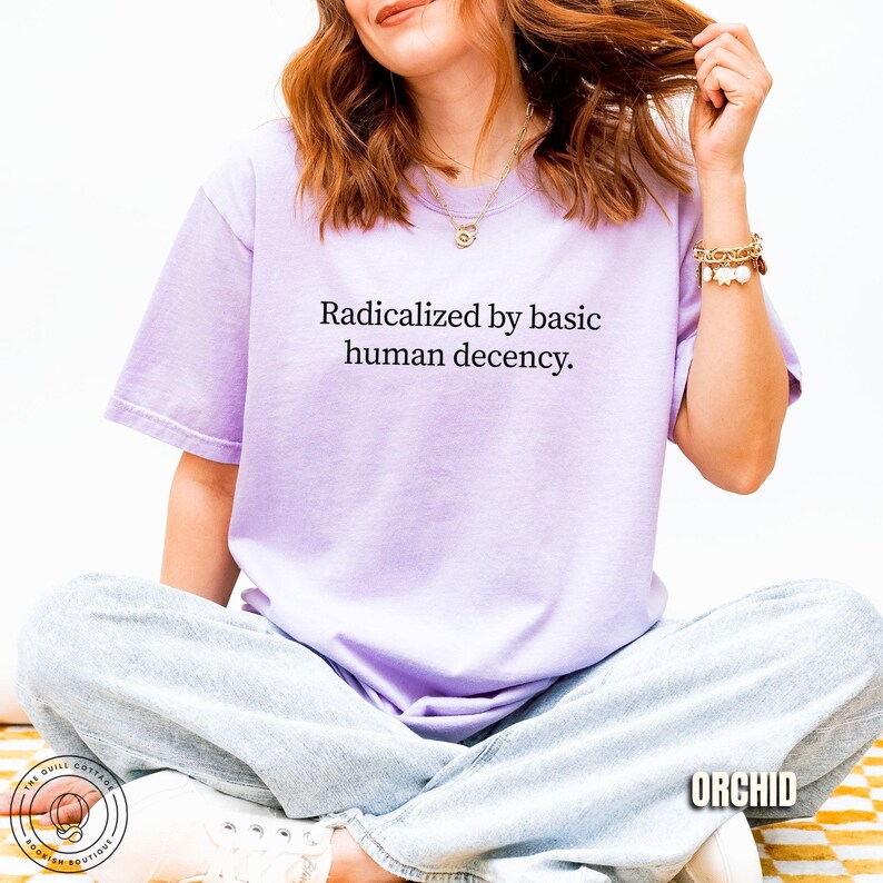 May include: A light purple t-shirt with the text "Radicalized by basic human decency." The shirt is a soft, comfortable material. The word "ORCHID" is printed on the bottom right corner of the image.