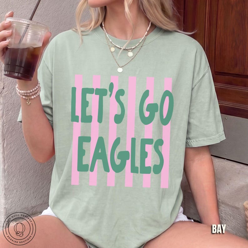 May include: A sage green short-sleeve t-shirt featuring a graphic design of vertical light pink stripes. The text "LET'S GO EAGLES" is printed in a bold, dark green, rounded font across the center of the shirt, layered over the striped pattern.