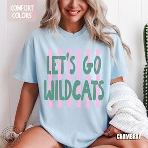 May include: A light blue short-sleeve t-shirt featuring vertical pink stripes and the green text "LET'S GO WILDCATS" printed on the front. The shirt has a crew neckline and a relaxed fit, with a small "COMFORT COLORS" label visible at the top left.