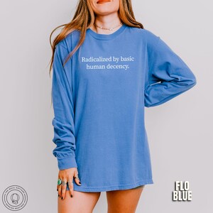 May include: A long-sleeved, Flo Blue shirt with the text "Radicalized by basic human decency" in white. The shirt is a solid color and has a relaxed fit. The Quill Cottage Boutique logo is visible in the bottom left corner.
