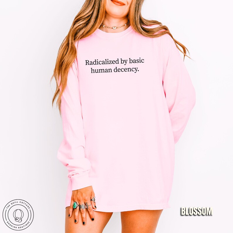 May include: A light pink long-sleeve shirt with the text "Radicalized by basic human decency" in black. The shirt has a crew neck and long sleeves. The word "Blossom" is printed in a black, bold font on the bottom right.