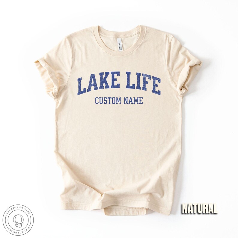Peut inclure: Un t-shirt cr&egrave;me avec les mots "LAKE LIFE" en lettres bleues et "CUSTOM NAME" en dessous. Le t-shirt est &eacute;tiquet&eacute; "NATURAL" en police de caract&egrave;res. Le logo Quill Cottage est dans le coin inf&eacute;rieur gauche.