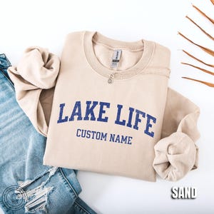 May include: A sand-colored crewneck sweatshirt featuring the blue text "LAKE LIFE" in a distressed font with the words "CUSTOM NAME" printed underneath. The sweatshirt is paired with blue denim jeans that have distressed detailing on the leg.