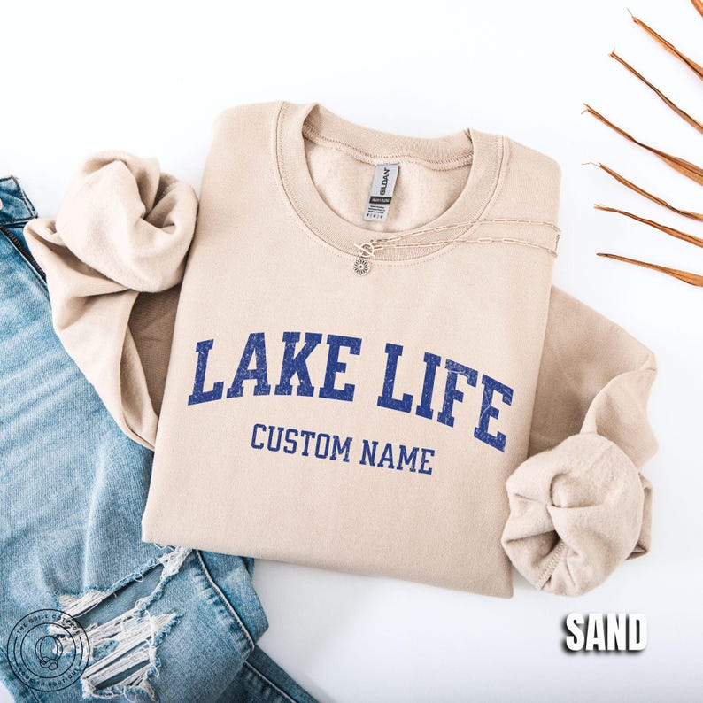 May include: A sand-colored crewneck sweatshirt featuring the blue text "LAKE LIFE" in a distressed font with the words "CUSTOM NAME" printed underneath. The sweatshirt is paired with blue denim jeans that have distressed detailing on the leg.