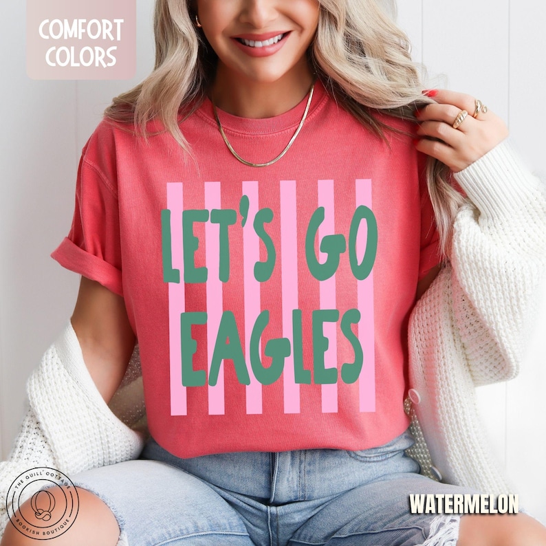 May include: A coral red short-sleeved t-shirt featuring vertical light pink stripes and the dark green text "LET'S GO EAGLES" centered on the front. The shirt has a crew neckline and a relaxed fit, with a small "COMFORT COLORS" label visible.