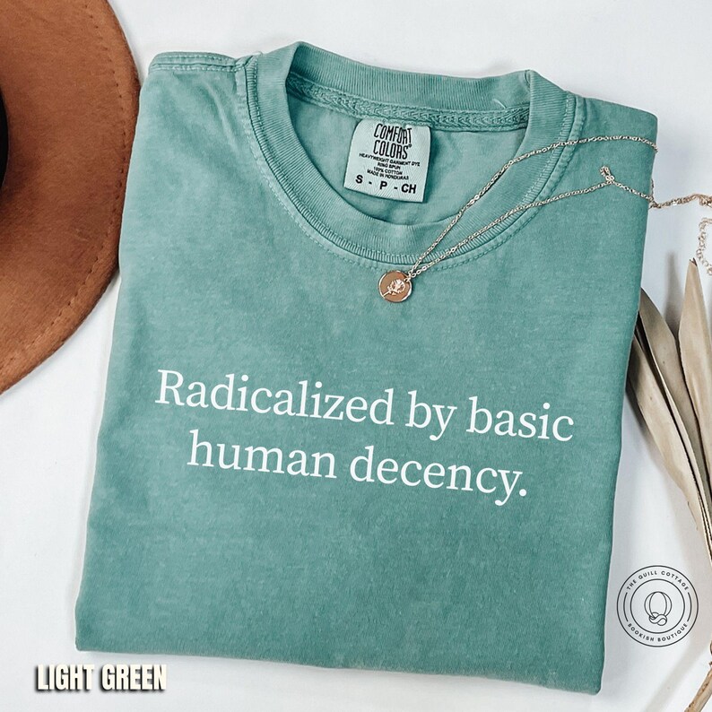May include: Light green t-shirt with the text "Radicalized by basic human decency" in white. A gold-colored necklace with a pendant rests on the shirt. The shirt is from Comfort Colors and is size small.