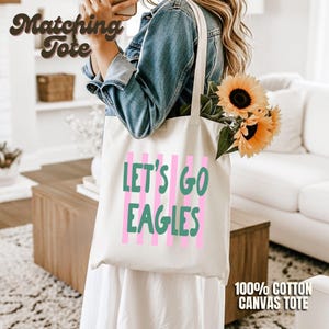 May include: A cream-colored canvas tote bag featuring vertical pink stripes and the green text "LET'S GO EAGLES" printed on the front. The bag is carried over the shoulder and contains a bouquet of yellow sunflowers with green stems and leaves.