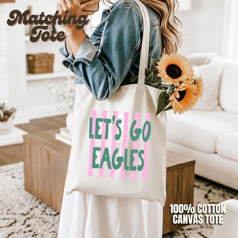 May include: A cream-colored canvas tote bag featuring vertical pink stripes and the green text "LET'S GO EAGLES" printed on the front. The bag is carried over the shoulder and contains a bouquet of yellow sunflowers with green stems and leaves.