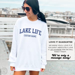 May include: A white long-sleeve sweatshirt featuring the dark blue text LAKE LIFE and CUSTOM NAME printed on the chest. The garment has a crew neckline and a relaxed fit. A white box with black text reads LOVE IT GUARANTEE and We're only a message away!