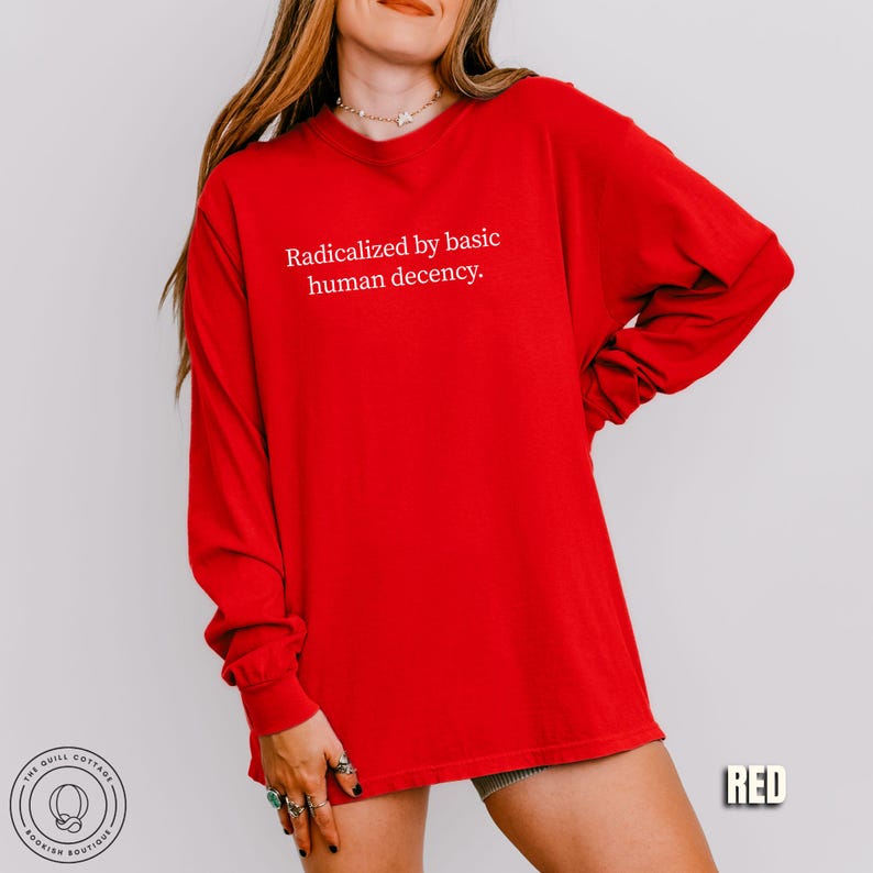 May include: A bright red long-sleeve shirt with the text "Radicalized by basic human decency." in white. The shirt is a solid color and appears to be made of a soft material. The image also includes a small logo in the bottom left corner and the word "RED" in the bottom right corner.