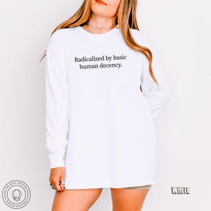 May include: White long-sleeve shirt with the text "Radicalized by basic human decency." The shirt has a crew neck and a relaxed fit. The Quill Cottage logo is visible on the bottom left. The word "WHITE" is on the bottom right.