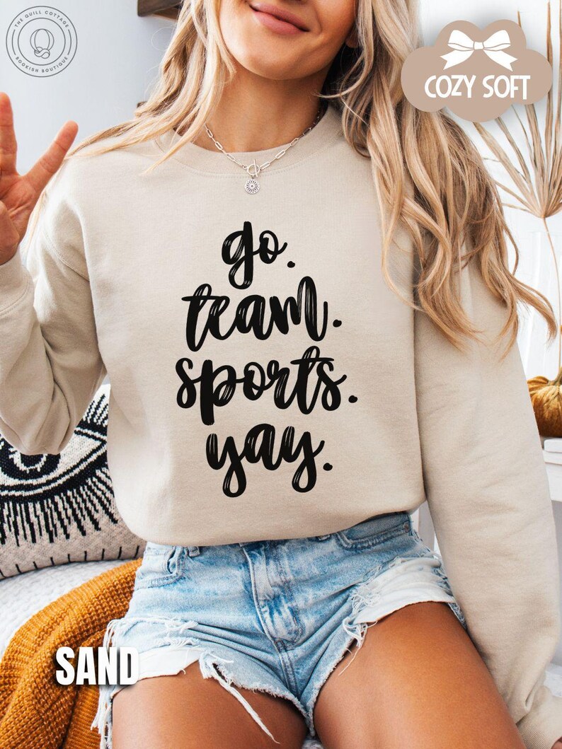 May include: A beige sweatshirt with the text 'go. team. sports. yay.' in black lettering. The sweatshirt is worn by a person wearing denim shorts. The text 'COZY SOFT' is in a white cloud with a pink bow on top of the cloud. The text 'SAND' is on the bottom left of the image.