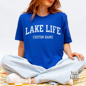 Peut inclure: T-shirt bleu roi avec l'inscription "LAKE LIFE" en lettres blanches au-dessus de "CUSTOM NAME". Le t-shirt est &agrave; manches courtes et a un col rond. L'image montre &eacute;galement un jean bleu clair.