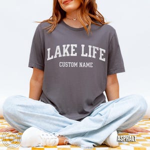 Peut inclure: T-shirt gris asphalte avec le texte "LAKE LIFE" en lettres blanches. En dessous, le texte "CUSTOM NAME". Le mod&egrave;le porte un jean bleu clair et des baskets blanches. Le t-shirt est de "The Quiet Bookish Boutique".